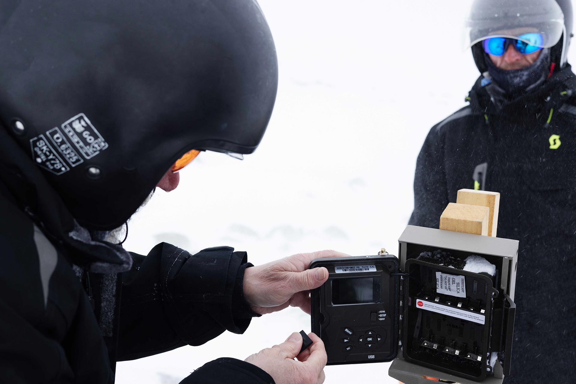 Two people in snow gear updating the memory card of the camera attached to the feeding station in the Arctic tundra